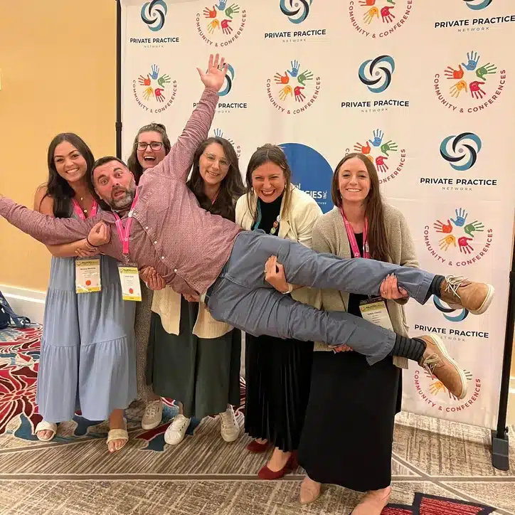Six people smiling and posing together, with one person being carried horizontally by the others in front of a branded conference backdrop.