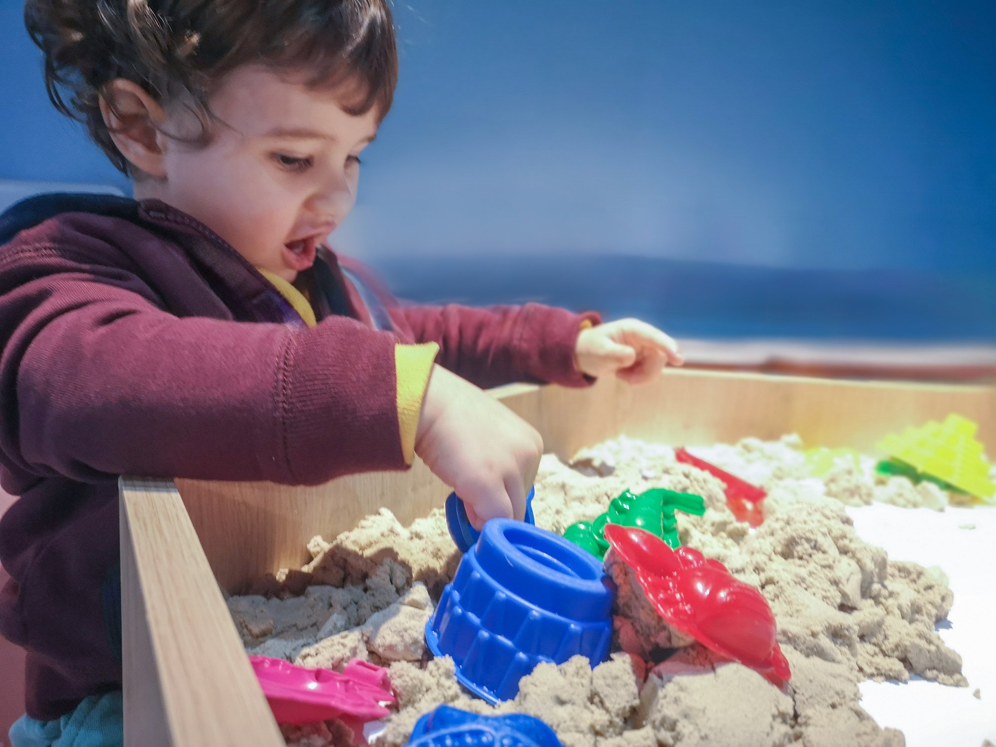 A young child wearing a maroon hoodie plays with colorful plastic toys in a sand table, engaging in imaginative play, set against a gradient blue background.